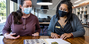 Two women wearing masks sitting at a table talking and looking at photographic slides