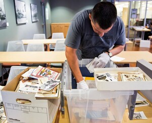 A man wearing white cotton gloves sorts through photos and documents from a bin.