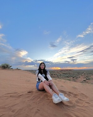 Photo of Myacedes Miller sitting on a hill composed of tan sand with footprints around her. She is sitting in front of a sunset wearing white shoes and jean shorts. The sky is blue with clouds blotting an orange sun low on the horizon.