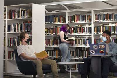 Three students in the library stacks talking looking at books