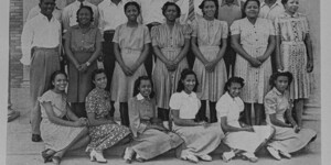 An archival black and white image of a group of people posed for the camera sitting in front and standing in back