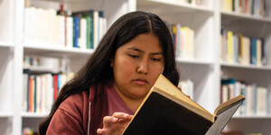 An ASU Library student worker reads a book in the newly renovated Hayden Library, sitting in front of white bookshelves filled with books.