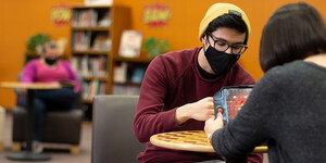 Two students play a game while wearing masks, another person reads by themselves in the background.