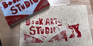 A linocut stamp of a block and print with Book Arts Studio text in red ink