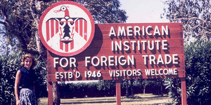 Student standing next to the original welcome sign for the American Institute for Foreign Trade, which reads “established 1946, visitors welcome”.