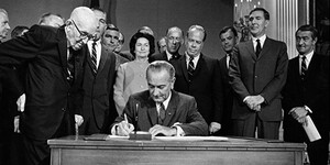 President Lyndon Johnson signs Central Arizona Project legislation seated at a ceremonial desk, while advisors and the First Lady Lady Bird Johnson stand behind him.