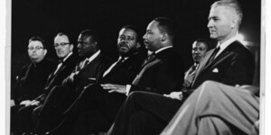 Black and white archival photo of Martin Luther King Jr. sitting with a group of people on stage