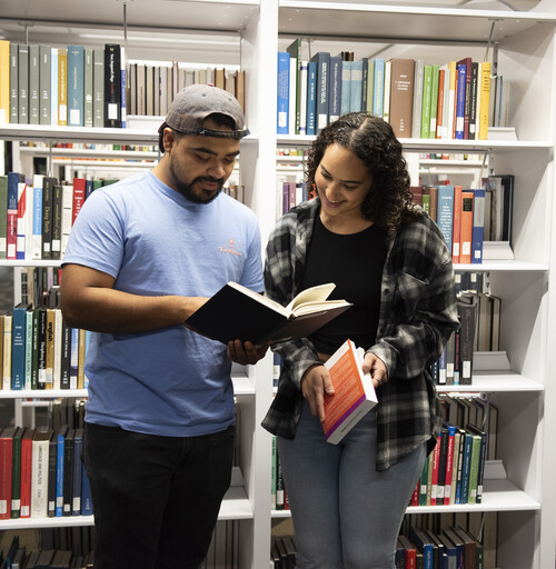 Two students enjoy reading a book in front of library bookshelves.