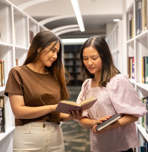 Two students look into an open book together in library bookshelves.