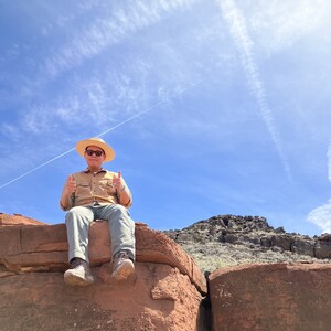 Photo of Eli Shepherd (Dine') sitting on large red rocks wearing a hat in front of blue skies