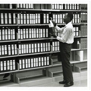 A man examines a microcard document pulled from a wall of shelves filled with microcard boxes.
