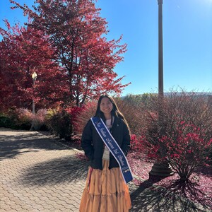 Photo of Tonana Ben, Library Aide at Labriola Center. She is standing in front of red bushes and a red tree wearing a long yellow skirt with a sash across her shoulder that says "Miss Indian Arizona 2nd attendent" 