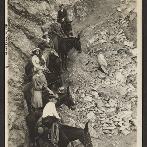 Black and white print of five women and one man riding mules on the Bright Angel Trail. A wooden sign next to the trail says, "Photo by Kolb Bros." Handwritten annotation, "Annie Goodenough on Bright Angel Trail".