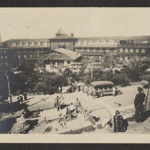 Black and white postcard with front elevated view of touring cars and visitors. Native American individuals and crafts are visible near entrance.