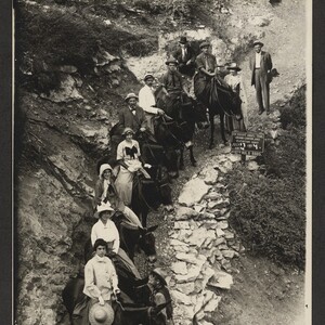 Black and white print with members of the Luhrs family.  Typescript annotation that reads,"July 17th, 1914. Going down Bright Angel Canyon of Arizona, Those on the mules, from bottom up: _____, _____, Ella Luhrs, Emma Luhrs, George H. N. Luhrs, _____, Roger Hunt, George Luhrs Jr., Catherine Margarita "Gretchen" (Mrs Geo H. N.) Luhrs standing beside George Luhrs Jr. Featured in the picture is a sign that reads "Photo by Kolb Brothers".