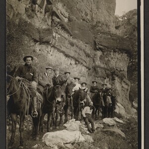 Black and white print of six men and four women (unidentified) astride mules on a trail. One man sits on rocks beside the trail.