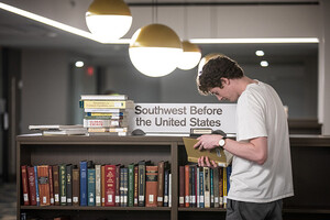 Man standing in front of a book shelf looking at an open book he is holding in his hands.