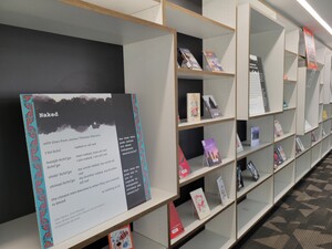 A multi-colored cardboard sign with a poem printed on it, surrounded by shelves containing a variety of Indigenous Poetry books. 