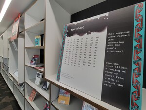 A multi-colored cardboard sign with a poem printed on it, surrounded by shelves containing a variety of Indigenous Poetry books. 