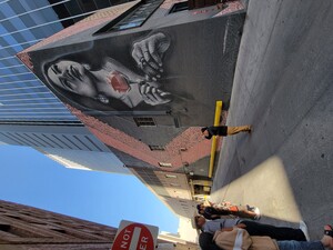 Photo of Breeze speaking in front of the second mural in downtown Phoenix. This mural is a black and white portrait of a young girl from the community holding a red rose looking East. The black and white and gray portrait contains spirals beneath, which weave throughout the mural and can be seen in the triangular border on either side of the girl.