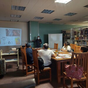 Pictured is Kayla Shaggy presenting on her experience of being an Indigenous zine and comic book artist. The projector shows her featured in a film called, "Unsolved Mysteries". An Indigenous family of four are a part of the workshop's audience along with another Indigenous student.  