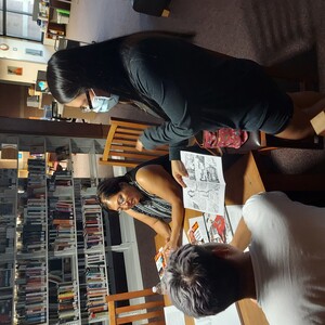 Pictured is Kayla Shaggy showing a copy of her zine, "Godzilla Decolonizes Durango!" to an Indigenous student from Mesa Community College looking to transfer to Arizona State University. The student's family member is to the right of Kayla Shaggy. Behind Kayla and the audience is the Open Stacks and Distinctive Collection.