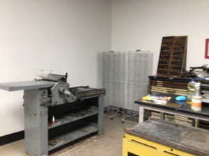 Photo of Jacob Meders' print shop in the University Center Building, West Valley - UCBB119. A large metal printing press is on the right. There are cases of metal letters held in sturdy wooden drawers to the right.