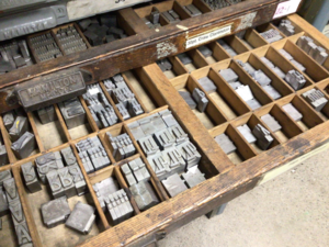 Photo of one of the wooden cases with various metal letters. The letters are heavy. The reason why we say "upper case" and "lower case" is because letters such as these were stored in cases, where the capital letters are stored in the upper case due to not being used as much as the lower case letters, which were stored in the more accessible cases for newsprints. 