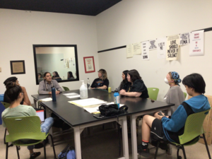 Photo of students sitting around a black table brainstorming three words to print with the printing press. Professor Meders sits at one end of the table.