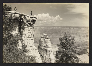 Black and white photo of a person with a camera and a park ranger overlooking the Grand Canyon from the North Rim. Trees are in the canyon walls and puffy white clouds are seen above the canyon.