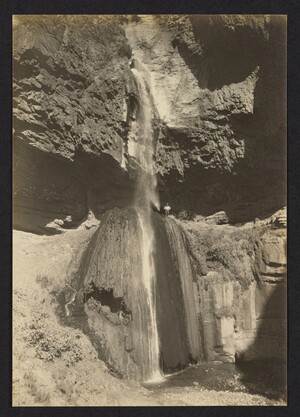 Black and white photograph of a person standing next to a very tall two stage waterfall with a large cave opening behind him in the canyon wall.