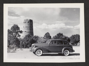 1930's automobile with white wall tires and a male driver in a hat behind the wheel and parked in front of the Hopi Watchtower.