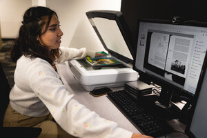 Person sitting at a desk looking at a desktop computer while scanning printed materials on a flatbed scanner