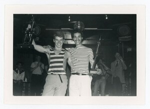  Two men with their arms around each other smile at the camera while holding trophies in their free hands. 