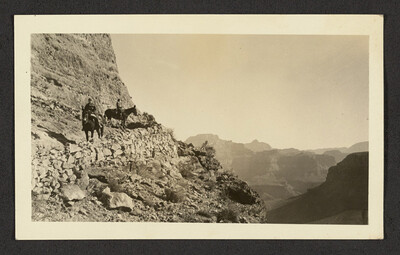 Black and white photograph depicting two visitors riding mules on the trail and looking into the canyon. The edge of the trail is reinforced with loose stones and a beautiful canyon vista is seen in the distance.
