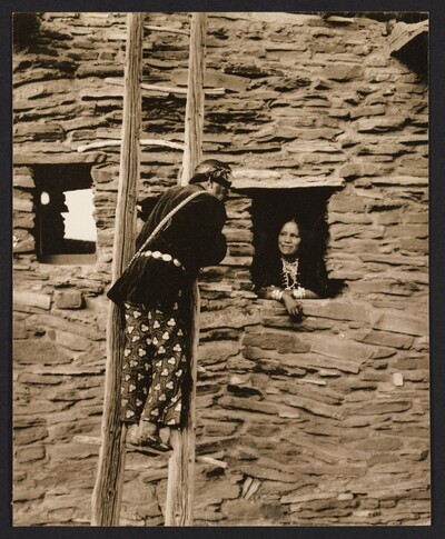 Black and white photograph of a person on a handmade wooden ladder climbing up the wall of a tall adobe structure with window openings (possibly the Hopi Watchtower) with another person leaning out of the window opening.