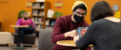 Two students play a game while wearing masks, another person reads by themselves in the background.