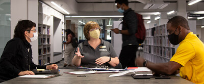 Three people wearing masks talk together at a table with piles of papers on it; students browse books behind them.