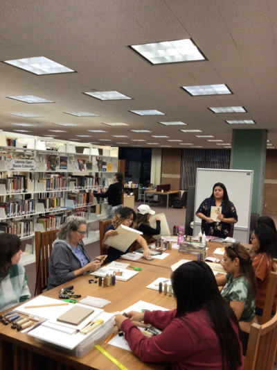 Photos of Vina Begay (Diné) at the front of a large wooden table with participants seated. The Labriola Center's open stacks are to the left of the photo. Paper, scissors, and kits are in front of participants. Vina is teaching participants how to bind their books. 