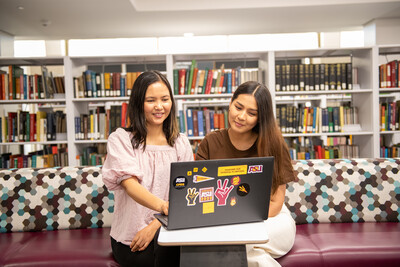 Two people sitting in the library looking at a laptop together with shelves of books behind them