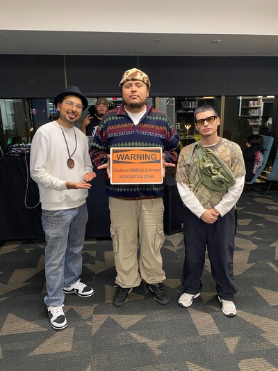 Pictured from left to right: Director Alex Soto (Tohono O'odham), DJ Ishaboi (O’otham/Diné), and DJ AlexCvstles (O’odham) holding an orange warning sign that states, "O'odham HIMDAG Enforced INDIGENOUS ZONE." Dj Acro (Hopi) is throwing a peace sign in the background behind DJ Ishaboi and Director Alex Soto.