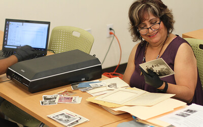 A woman wearing archival gloves smiles at old photos of her family while getting ready to use the scanner on the table in front of her.