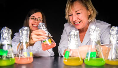 Two laughing scientists inspect beakers filled with pink, green, and yellow liquid.