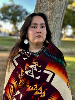 Mirra Keeto (Diné / Akimel O’odham) wearing a cream and sunset color blanket over shoulders, turquoise jewelry, and round shell earrings.