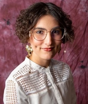 A person with curly hair and glasses smiles at the camera, wearing a white lace-detailed top.